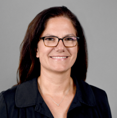 Melanie Gall headshot - fair-skinned woman, smiling, brown hair, glasses, black collared shirt, necklace